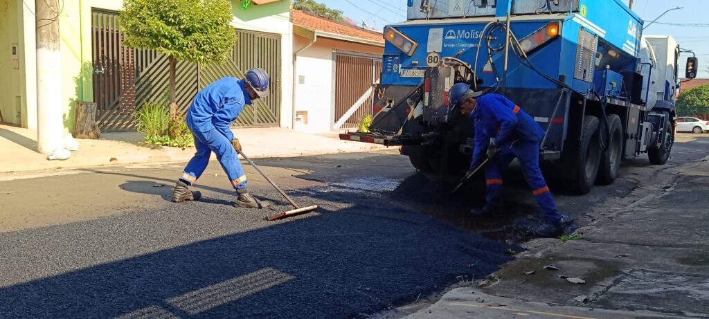 Nesta imagem, vemos dois operários realizando um serviço de manutenção ou pavimentação em uma rua: Ação: Eles estão espalhando massa asfáltica fresca sobre o solo. Um trabalhador, à esquerda, usa um rodo largo para nivelar o asfalto, enquanto o outro, à direita, usa uma ferramenta manual para auxiliar no processo próximo a um caminhão. Equipamento: Há um grande caminhão azul da empresa "Molise Serviços e Construções Ltda" estacionado, que parece ser um equipamento especializado para a distribuição de asfalto. Ambiente: O cenário é uma rua residencial em um dia ensolarado. Ao fundo, percebem-se casas com portões de metal e uma árvore. Os operários usam uniformes azuis e equipamentos de segurança, como capacetes.