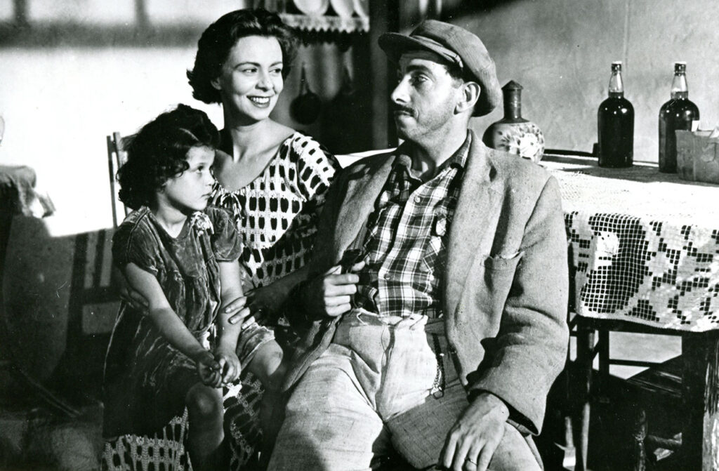 Esta fotografia clássica em preto e branco captura um momento íntimo de uma família simples em um ambiente doméstico rústico, evocando um sentimento de conexão emocional e vida cotidiana A composição da cena é organizada com os três personagens centralizados, criando uma sensação de unidade e proximidade. O homem encontra-se em primeiro plano, ocupando o lado direito, enquanto a mulher e a criança estão sentadas mais ao fundo, à esquerda, formando um triângulo visual equilibrado. A interação é marcada pelo olhar da mulher para o homem, sugerindo uma cumplicidade silenciosa, enquanto a criança olha para frente, conferindo uma profundidade narrativa que sugere uma cena capturada de um filme ou peça de época. O homem é o foco central, vestindo uma jaqueta sobre uma camisa xadrez e um boné, com um bigode proeminente que lhe confere um ar de seriedade e maturidade. A mulher, ao lado, possui cabelos escuros e ondulados, expressando um sorriso suave e sereno enquanto olha para o homem. Ela veste uma blusa com um padrão geométrico distinto. A criança, uma menina com cabelos encaracolados e vestida com um traje simples e escuro, está sentada no colo ou ao lado da mulher, exibindo uma expressão contemplativa e observadora que contrasta com a calma dos adultos. A imagem é um exemplo notável da fotografia cinematográfica clássica, possivelmente do cinema neorrealista, caracterizada por um forte contraste entre luz e sombra que define os volumes e as texturas. A técnica utiliza iluminação lateral dramática, que realça os traços faciais dos sujeitos e adiciona profundidade ao ambiente, conferindo uma qualidade atemporal e autêntica à obra. O foco é nítido nas expressões, enquanto os detalhes do ambiente permanecem secundários, priorizando a conexão humana. O cenário parece ser o interior de uma casa humilde, com uma mesa coberta por uma toalha de crochê detalhada, onde repousam garrafas que remetem a um ambiente rural ou de época. A luz que incide lateralmente cria sombras longas e definidas na parede branca atrás dos sujeitos, reforçando a textura da cena. Essa iluminação cria um contraste marcante, envolvendo os personagens em uma atmosfera de nostalgia e simplicidade, onde o ambiente doméstico serve como um reflexo da vida cotidiana e dos laços afetivos que unem a família representada.