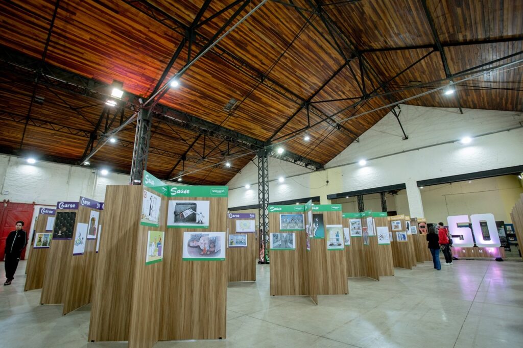 A imagem mostra um grande salão de exposições, com teto alto revestido de madeira e estruturas metálicas aparentes. Painéis de madeira organizam diferentes estandes, decorados com ilustrações e textos nas cores verde e roxa, com temas como “Saúde” e “Carga”. Algumas pessoas caminham pelo espaço, enquanto, ao fundo, há uma instalação iluminada com letras grandes.