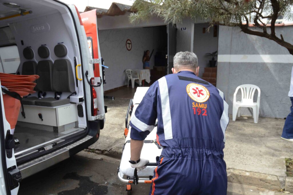 A imagem mostra uma equipe de atendimento de emergência ao lado de uma ambulância com as portas traseiras abertas. No interior, há uma maca, equipamentos médicos e assentos. À direita, uma profissional usando uniforme azul-marinho com faixas refletivas laranja segura uma maca ou cadeira de transporte. A cena parece ocorrer ao ar livre, em frente a um prédio, durante o dia.