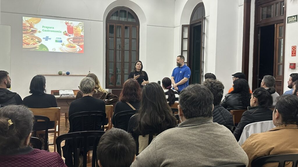 A imagem mostra uma palestra ou reunião sobre gastronomia em uma sala ampla, com janelas altas e portas de madeira. Uma mulher fala diante do público, acompanhada por um homem usando uma camiseta azul. As pessoas estão sentadas em cadeiras, voltadas para os palestrantes. No projetor, aparece a apresentação “Prepara Gastronomia”, com os logotipos do Sebrae e da PIRA, além de imagens de hambúrgueres, massas, bebidas e sobremesas. Na porta, há uma placa indicando “Saída de emergência”.