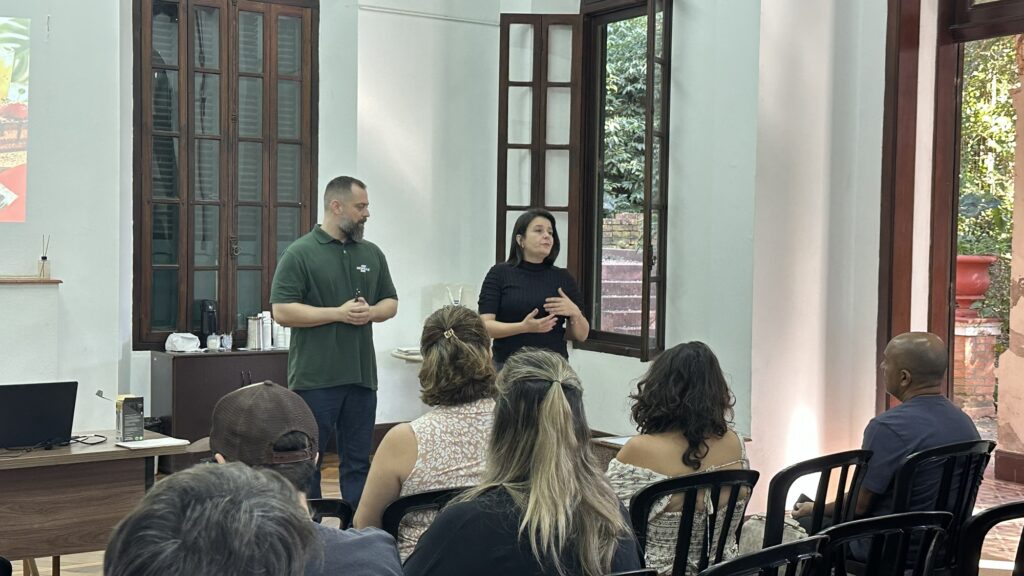 Um homem e uma mulher estão apresentando para um grupo de pessoas sentadas, com uma janela ao fundo. A cena se passa em uma sala com paredes brancas e detalhes em madeira escura. Há duas grandes janelas de madeira com venezianas, uma delas aberta, revelando vegetação e uma escada externa. No lado esquerdo da sala, uma mesa de madeira abriga um laptop preto, óculos, e uma caixa de produto. Atrás da mesa, um armário escuro de madeira está posicionado sob uma janela fechada com múltiplas divisões. Sobre o armário, há um dispensador de ar, uma cafeteira, e recipientes metálicos. Uma projeção em uma parede à esquerda mostra imagens coloridas, possivelmente de alimentos ou produtos. No centro da imagem, em pé, um homem com barba grisalha veste uma polo verde escura com um logo branco e calça jeans, segurando um pequeno objeto em suas mãos. Ao seu lado direito, uma mulher com cabelos escuros usa uma blusa preta de gola alta e gesticula com as mãos enquanto fala, olhando para a audiência. Sentadas em cadeiras pretas em primeiro plano e em segundo plano, estão várias pessoas, a maioria mulheres, com diferentes penteados e roupas. Algumas estão viradas para os apresentadores, enquanto outras têm suas costas voltadas para a câmera. A iluminação na sala parece ser uma combinação de luz natural vinda das janelas e iluminação artificial, criando um ambiente de aprendizado ou apresentação. A atmosfera é profissional e focada, com a audiência prestando atenção aos palestrantes. As cores predominantes são branco nas paredes, madeira escura nos móveis e janelas, verde na polo do homem e tons variados de roupas e cabelos nas pessoas.