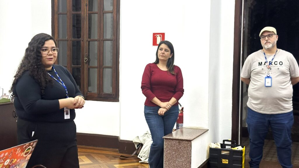 Vejo três pessoas de pé em uma sala interna, aparentemente durante uma apresentação ou conversa: À esquerda: mulher de pele clara, cabelos longos e cacheados, usa óculos e roupa escura (blusa preta). Está falando ou explicando algo, com as mãos à frente; usa crachá pendurado no pescoço. Ao centro: mulher de blusa vinho/malha e jeans, em pé encostada numa mureta baixa, braços relaxados à frente; expressão séria/atenta. À direita: homem com boné claro, óculos e barba curta, veste camiseta clara com a palavra “MARCO” impressa e também usa crachá; está com as mãos atrás das costas. Ambiente: parede branca com janela de madeira ao fundo à esquerda e um extintor/sinal de emergência visível atrás da pessoa do centro; piso de madeira com padrão; à esquerda, na parte inferior da imagem, nota-se um laptop com adesivos; no canto direito inferior há caixas/caixa de ferramentas pretas e amarelas. Iluminação: interna, clara e uniforme, sem sombras marcantes. Atmosfera: encontro informal ou reunião — alguém fala enquanto os outros escutam/observam.