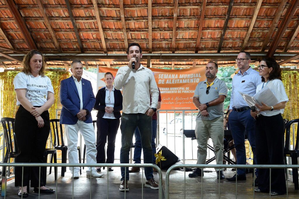 Esta foto mostra um grupo de pessoas em um palco ou plataforma durante um evento oficial. Aqui estão os principais detalhes: O Grupo: Há sete pessoas em pé em uma linha. A pessoa central, um homem de camisa clara e calça jeans, está segurando um microfone e falando. Ele está posicionado como o orador principal. O Evento: Ao fundo, um banner laranja estendido indica que se trata da "Semana Mundial de Aleitamento" e da "Semana Municipal de Aleitamento de Piracicaba". Também é mencionado o "Comitê de Aleitamento Materno e Alimentação Complementar". Vestimenta: As pessoas vestem roupas casuais e sociais. Duas mulheres, uma na extrema esquerda e outra na extrema direita, vestem camisetas brancas com dizeres sobre aleitamento, indicando que fazem parte da organização ou apoio ao evento. Ambiente: O local possui um teto de telhas de barro e vigas de madeira aparentes, com cortinas douradas decorativas ao fundo, criando um ambiente que parece ser um pavilhão ou espaço de eventos comunitários. Há também grades metálicas protegendo a frente do palco.