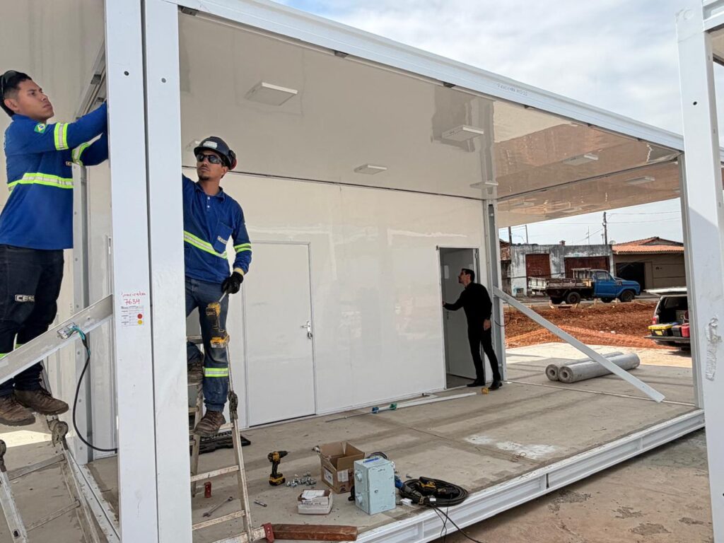 Esta fotografia captura uma cena de trabalho em um canteiro de obras, onde dois profissionais realizam a montagem e instalação de uma estrutura modular metálica ao ar livre. A composição da cena é organizada em torno da estrutura de metal branca, que atua como uma moldura natural para a ação. Dois trabalhadores estão posicionados em primeiro plano na parte frontal e lateral, focados na montagem da viga, enquanto um terceiro homem aparece ao fundo, à direita, entrando ou saindo por uma das portas da estrutura. A disposição dos elementos destaca o processo de construção, com ferramentas, caixas e materiais espalhados pelo chão de concreto, sugerindo um ambiente de trabalho ativo e em progresso sob condições de luz natural. Os dois trabalhadores principais estão uniformizados com camisas de manga longa azuis, faixas refletivas de segurança, calças jeans e botas robustas, sendo que ambos utilizam equipamentos de proteção individual, incluindo capacetes e óculos de segurança. O homem à esquerda está em uma posição elevada, utilizando uma escada, e parece estar fixando partes da estrutura superior, enquanto o seu colega, também em uma escada, opera uma ferramenta elétrica com foco total. O terceiro homem ao fundo veste roupas escuras e mantém uma postura mais casual, observando ou interagindo com a entrada da unidade modular. A imagem é uma fotografia documental realista, caracterizada pela clareza técnica e pelo registro fiel de um ambiente de trabalho industrial. Não há filtros artísticos estilizados, focando-se na nitidez dos detalhes, desde a textura metálica da estrutura até as ferramentas espalhadas no chão. A perspectiva é frontal e ligeiramente elevada, o que permite uma visualização abrangente tanto do detalhamento da montagem quanto do cenário ao redor, mantendo o foco nítido em todas as profundidades da imagem. O cenário é um terreno aberto, possivelmente um lote em desenvolvimento, sob um céu nublado que proporciona uma iluminação difusa e uniforme, eliminando sombras fortes e permitindo que todos os detalhes sejam visíveis. A atmosfera é de produtividade e foco profissional, com elementos como o solo de terra batida ao fundo e o entorno urbano simples que contextualizam a natureza prática da tarefa. A luz suave do dia contribui para um clima sóbrio, realçando as superfícies reflexivas da estrutura branca e os detalhes das ferramentas, criando um ambiente funcional e típico de uma obra em execução.