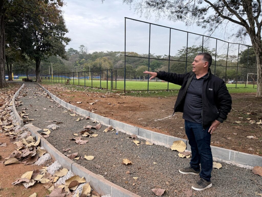 A imagem mostra um homem em pé, usando jaqueta preta, camisa escura e calça jeans, apontando para uma área de um parque esportivo. Ao lado dele há uma trilha curva em construção, delimitada por blocos de concreto e preenchida com pedras britadas, coberta por folhas secas. Ao fundo, aparecem árvores, cercas altas e campos esportivos gramados, incluindo uma quadra com traves. O local é arborizado e o céu está nublado.