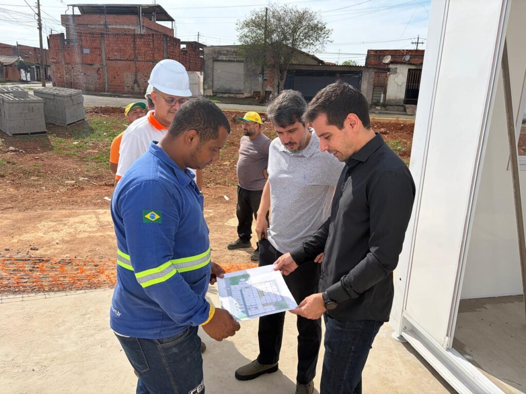 Uma cena de trabalho ao ar livre apresenta quatro homens em um canteiro de obras, focados na análise de plantas arquitetônicas enquanto discutem detalhes técnicos para o desenvolvimento do projeto local. A composição da cena é organizada de forma funcional, com os indivíduos agrupados no lado direito da imagem para examinar o documento impresso que seguram coletivamente. O enquadramento é em plano médio, permitindo observar a interação entre os participantes e o contexto da obra. O homem em primeiro plano, à esquerda, estabelece o ponto focal ao segurar a planta, enquanto os outros se inclinam em direção a ela. A disposição dos sujeitos cria um semicírculo informal, sugerindo uma colaboração profissional e uma comunicação direta entre a equipe técnica e administrativa. O grupo é composto por quatro homens. O primeiro, à esquerda, veste um uniforme de trabalho azul com faixas refletivas e uma bandeira do Brasil na manga. Atrás dele, outro homem usa um capacete branco e camisa clara. Ao centro, um terceiro homem utiliza uma camisa polo cinza, e à direita, um homem de camisa preta e calça jeans aponta para o papel. Suas expressões são de concentração e seriedade, típicas de uma análise técnica de engenharia. A postura corporal de todos indica engajamento com a tarefa em mãos, com inclinações atentas e olhares direcionados para o desenho impresso. Esta é uma fotografia digital documental, com uma estética limpa e luz natural direta. A execução técnica privilegia o foco nítido nos sujeitos principais e no objeto da interação, enquanto o fundo permanece com uma nitidez moderada que mantém o contexto geográfico identificável. A paleta de cores é composta por tons terrosos do solo, o azul vibrante do uniforme e o contraste entre as roupas claras e escuras dos outros profissionais, criando um equilíbrio visual que destaca os trabalhadores em relação ao ambiente industrial inacabado. O cenário é um canteiro de obras aberto, com solo de terra batida e blocos de construção empilhados no fundo, sob um céu claro e ensolarado. A iluminação é difusa, proveniente de um dia iluminado, o que reduz sombras agressivas e permite uma leitura clara de todos os elementos da imagem. O ambiente transmite uma atmosfera de produtividade, planejamento e desenvolvimento urbano, com a presença de construções de tijolos ao fundo que reforçam a natureza do local. O clima geral é de cooperação profissional, refletindo a rotina cotidiana de uma equipe de campo dedicada ao progresso de uma construção.