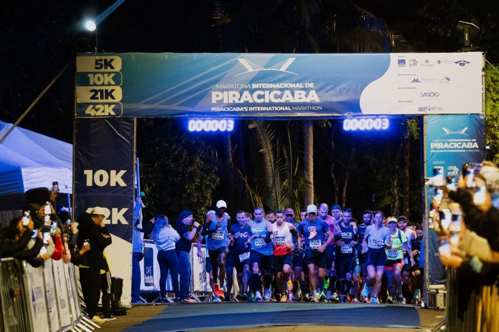 A imagem mostra a largada de uma corrida noturna da Maratona Internacional de Piracicaba. Diversos corredores avançam sob um pórtico iluminado, que indica as modalidades de 5K, 10K, 21K e 42K. O cronômetro marca poucos segundos após o início. O percurso é cercado por grades, tendas e espectadores que registram o momento com celulares. Ao fundo, há árvores e palmeiras, enquanto a iluminação azul destaca o evento.