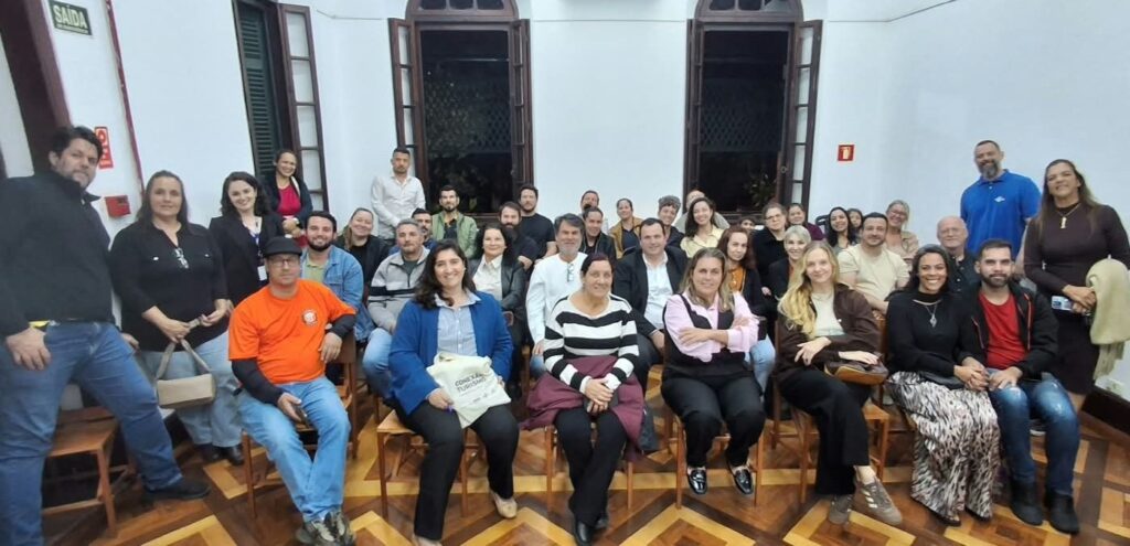 A imagem mostra um grupo grande de adultos reunido para uma foto em uma sala histórica. Algumas pessoas estão sentadas em cadeiras de madeira, enquanto outras permanecem em pé atrás delas. O ambiente tem paredes brancas, janelas altas de madeira, piso de parquet com desenho geométrico e sinalizações de segurança, incluindo “SAÍDA”. No centro, uma mulher segura uma sacola com a inscrição “Conselho de Turismo”. Todos parecem participar de uma reunião, evento ou atividade relacionada ao turismo.