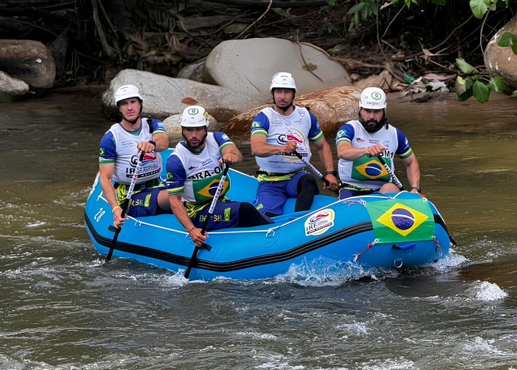 A imagem mostra quatro homens em um bote inflável azul, em movimento em um rio de águas correntes. Todos os homens estão usando capacetes de segurança brancos e uniformes com as cores do Brasil: azul, verde e amarelo. Os uniformes também exibem logotipos e textos relacionados a uma competição ou equipe. O bote possui bandeiras do Brasil em sua proa e em seu lado. Os homens estão segurando remos e parecem estar focados na atividade de rafting. A composição da imagem foca nos ocupantes do bote e na embarcação, com o fundo mostrando a vegetação natural e rochas às margens do rio. A atmosfera é de ação e competitividade, com um senso de trabalho em equipe e determinação. A iluminação sugere que é dia, com luz natural incidente sobre a cena.