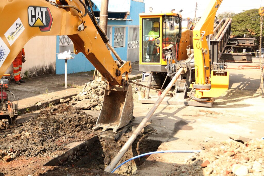 Uma escavadeira e um motorista trabalham em uma obra, escavando o chão para instalar um tubo. A imagem mostra uma cena de construção em um dia claro e ensolarado. Em primeiro plano, uma grande escavadeira amarela da marca CAT está posicionada ao lado de uma vala recém-aberta. A pá da escavadeira está abaixada e parece pronta para mover mais terra ou detritos. Um tubo longo e metálico atravessa a vala, parcialmente coberto por terra e cascalho. Mais ao fundo, a cabine da escavadeira é visível, e dentro dela, um motorista vestindo um capacete e um colete de alta visibilidade opera a máquina. À esquerda da escavadeira, um trabalhador com uniforme laranja de alta visibilidade está parado, usando luvas. Uma pequena máquina vibratória vermelha e cinza está posicionada no chão perto dele. Há também uma pilha de terra e entulho à direita da vala, indicando que o trabalho já está em andamento. Ao fundo, vê-se a fachada de uma casa ou edifício azul, com janelas e grades. O ambiente parece urbano, com calçada e um poste de luz ou fiação elétrica. A luz do sol é forte, criando sombras distintas no chão e nas máquinas. A atmosfera geral é de trabalho árduo e progresso de construção, com um clima de dia de trabalho eficiente.