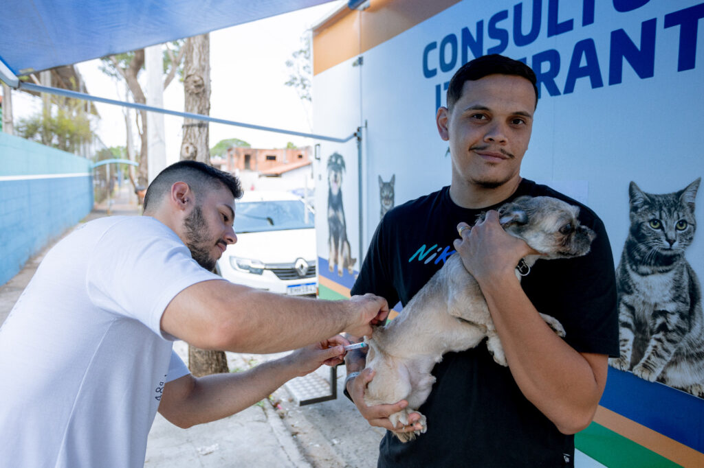 A imagem mostra duas pessoas em um espaço de atendimento veterinário itinerante. Uma delas segura com cuidado um pequeno cachorro, enquanto a outra aplica uma injeção no animal. Ao fundo, há uma parede com imagens de gatos e cães e o texto “CONSULTÓRIO ITINERANTE”, além de um veículo branco estacionado próximo.