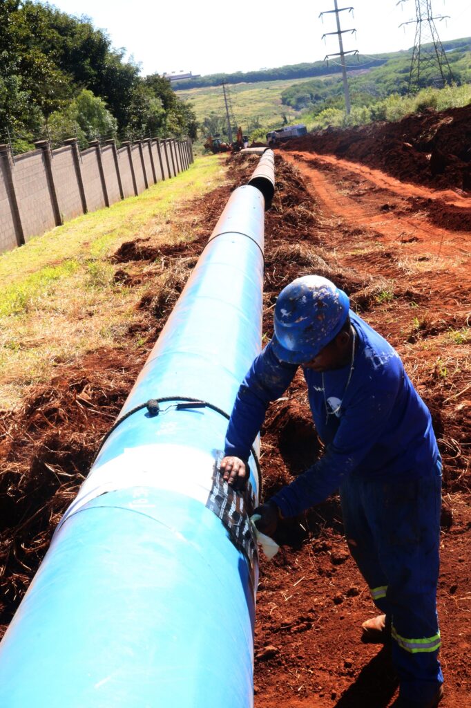 Uma vista em primeira pessoa de um trabalhador da construção civil em um canteiro de obras, colocando fita em um grande tubo azul. O trabalhador está vestindo um macacão azul e um capacete azul, e está ajoelhado ao lado do tubo. O tubo é grosso e azul, com algumas seções brancas e pretas de fita cobrindo ele. O terreno ao redor está coberto de terra vermelha e grama seca. Ao longe, é possível ver uma parede de concreto com arame farpado no topo, árvores verdes e colinas cobertas de vegetação verde, com linhas de energia ao fundo. O céu está limpo e branco, indicando um dia ensolarado. A composição coloca o trabalhador e o tubo em primeiro plano, ocupando a maior parte do quadro, e o cenário se estende ao fundo, criando uma sensação de profundidade e escala. As cores dominantes são o azul do tubo e do macacão, o vermelho da terra e o verde da vegetação. A imagem parece estar focada no trabalho do homem e na infraestrutura sendo construída.
