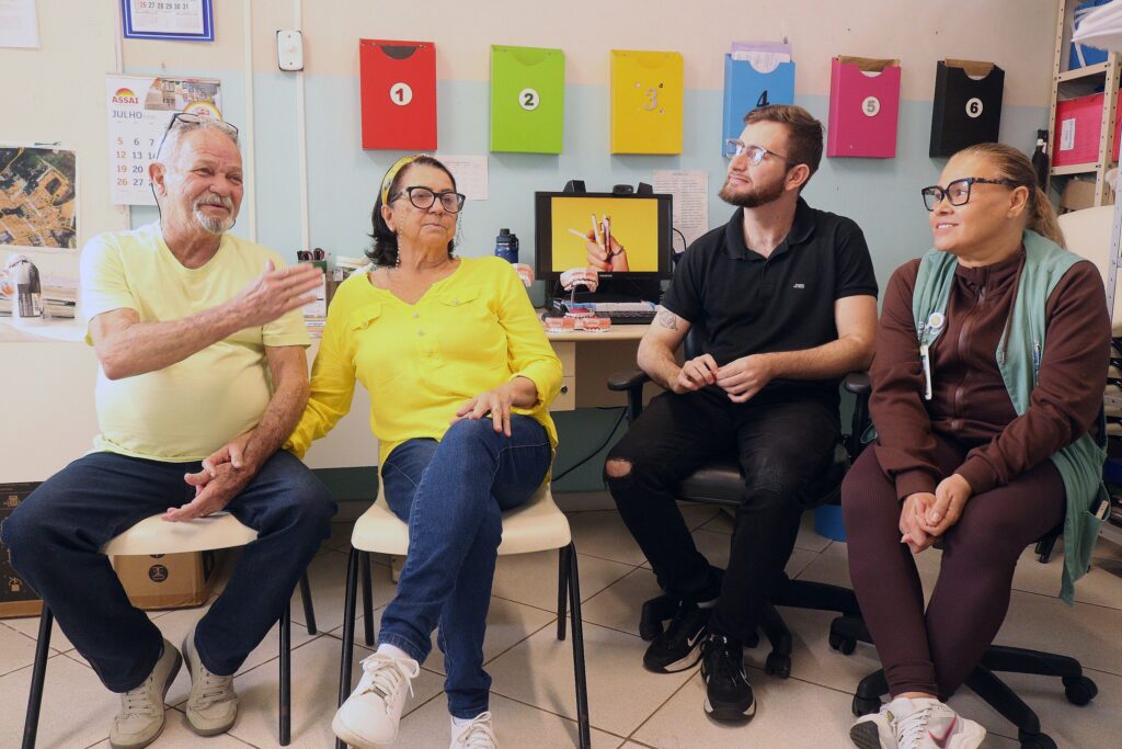 Cena em uma sala de atendimento ou reunião pequena, provavelmente em uma unidade de saúde ou clube comunitário. Quatro pessoas sentadas lado a lado formando um grupo: um homem idoso à esquerda, uma mulher idosa ao lado dele, um homem jovem com barba e óculos, e uma mulher adulta à direita. Todos parecem conversando de forma descontraída; o homem à esquerda gesticula enquanto fala, a mulher ao centro olha séria/atenta, o homem jovem sorri levemente olhando para a direita, e a mulher à direita sorri. Roupas e cores: duas pessoas usam tons amarelos (o homem e a mulher ao centro), o homem jovem veste preta e a mulher à direita usa marrom com colete verde-claro. Todos usam óculos. Ambiente: parede com organizadores coloridos (vermelho, verde, amarelo, azul, rosa, preto) numerados de 1 a 6; calendário e mapas pendurados; estante com pastas à direita. Mesa/mesa de trabalho ao fundo com um monitor mostrando uma imagem amarela, modelos de prótese/dentes, garrafa de água e materiais de escritório. Atmosfera geral acolhedora e comunitária, parece uma conversa em grupo ou consulta educativa (talvez sobre saúde bucal, pelo modelo de dentes visível).