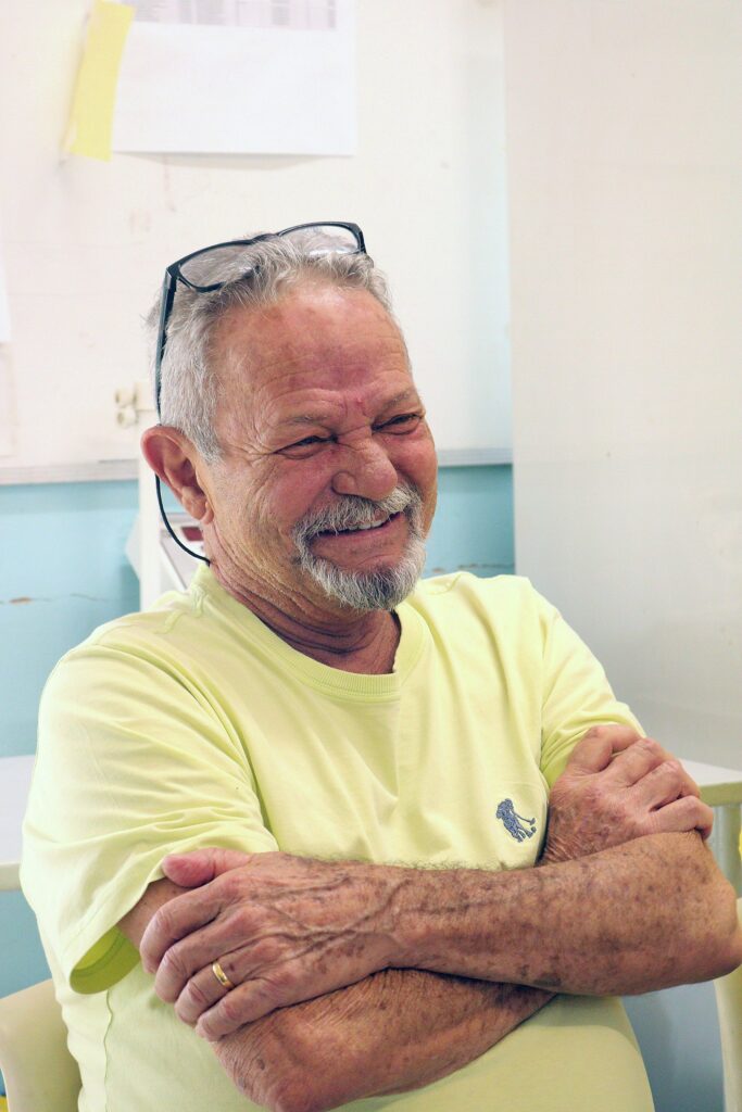 Homem idoso sentado, sorrindo com expressão alegre e olhos semi-fechados — parece rindo ou lembrando algo divertido. Detalhes físicos e roupa: Cabelos curtos e grisalhos; barba e bigode brancos com traços mais escuros. Rugas marcadas no rosto, principalmente ao redor dos olhos e testa. Usa óculos apoiados sobre a cabeça. Camiseta de tom amarelo claro com pequeno bordado azul no peito. Anel de casamento na mão esquerda. Postura e gesto: Braços cruzados sobre o peito, mãos apoiadas uma sobre a outra; postura relaxada e confortável. Sorriso amplo mostrando dentes; traços faciais indicam simpatia e leveza. Ambiente: Parede clara ao fundo com papéis presos e um quadro branco/armário sutilmente visível. Tom geral do ambiente é de interior, possivelmente sala de reunião, clínica ou espaço comunitário. Iluminação suave, predominância de tons claros e pastel no cenário. Impressão geral: Cena transmite aconchego, tranquilidade e convivialidade — a pessoa parece à vontade, participando de uma conversa informal.