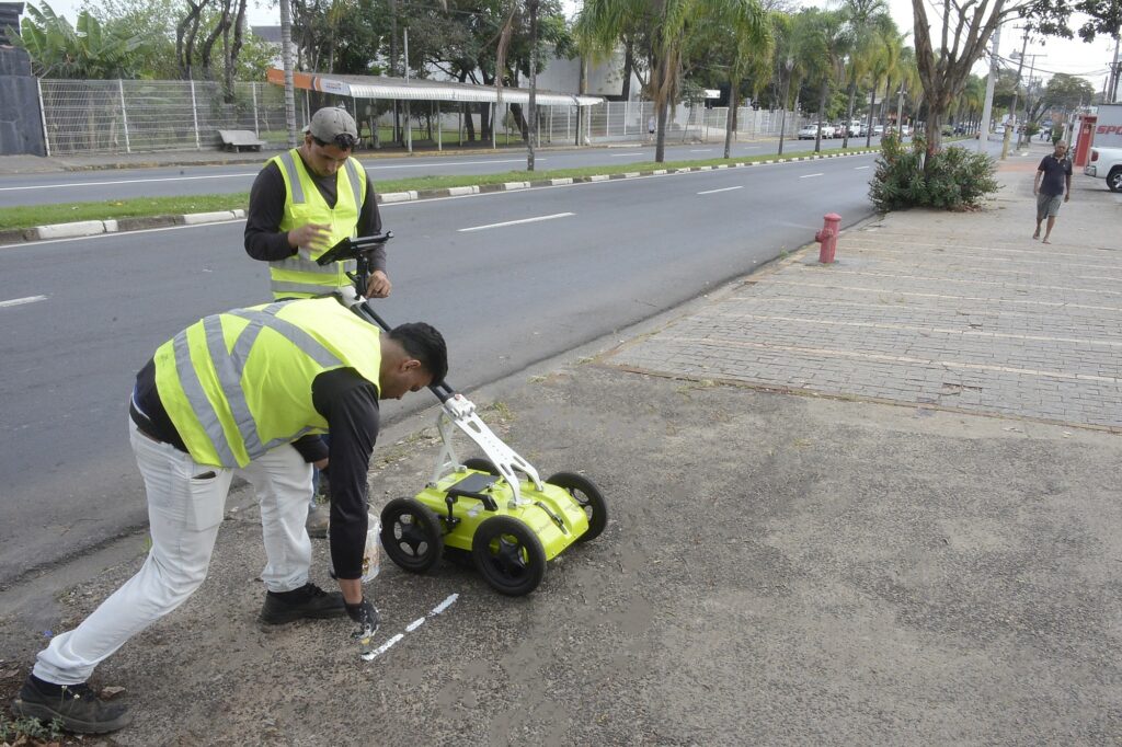 Na imagem vemos dois trabalhadores realizando uma atividade técnica na calçada/páreo da rua: Ambos usam coletes refletivos amarelos e roupas de trabalho; um está em pé manuseando um tablet acoplado a uma haste, o outro está curvado para frente, pintando ou marcando o solo com um pincel pequeno. Ao lado deles há um equipamento sobre rodas de cor verde-limão — parece um carrinho de varredura geofísica / radar de solo (GPR) ou instrumento de inspeção similar. Eles estão marcando uma linha branca no pavimento próximo à borda da calçada; ao redor nota-se asfalto e trechos de concreto com pequenas fissuras. A rua ao fundo é larga, com faixa central e canteiro gramado separando os sentidos; há palmeiras e um ponto de ônibus coberto no lado oposto. No passeio, à direita, há um hidrante vermelho e um homem caminhando mais adiante; carros estacionados aparecem ao longe. O dia está claro, iluminação natural, ambiente urbano com edificações e vegetação ao fundo.