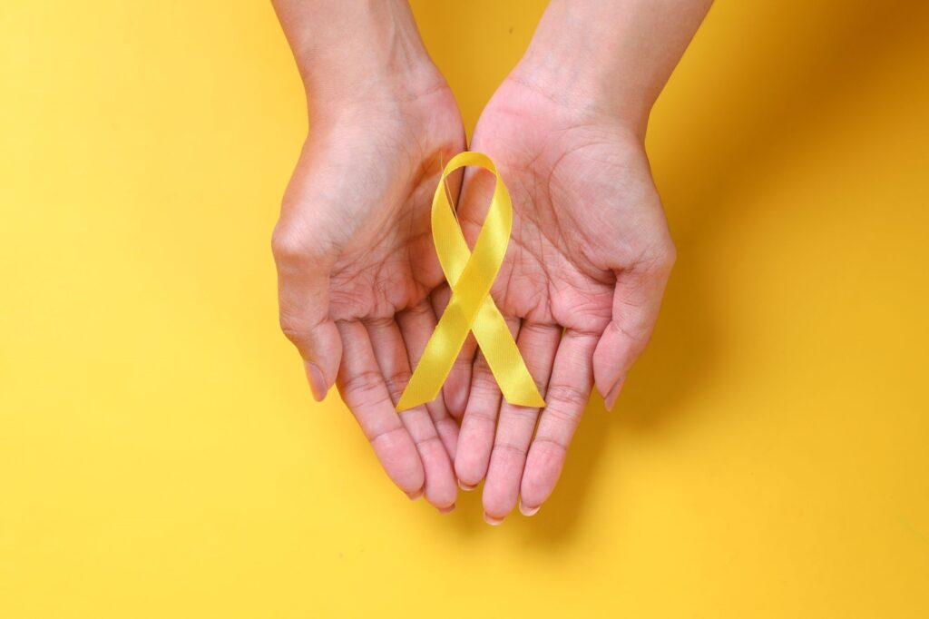A imagem mostra duas mãos abertas, uma sobre a outra, segurando uma fita amarela em formato de laço. O fundo é inteiramente amarelo, criando uma composição simples e vibrante. O laço amarelo costuma simbolizar campanhas de conscientização e prevenção, especialmente relacionadas ao suicídio e à saúde mental.