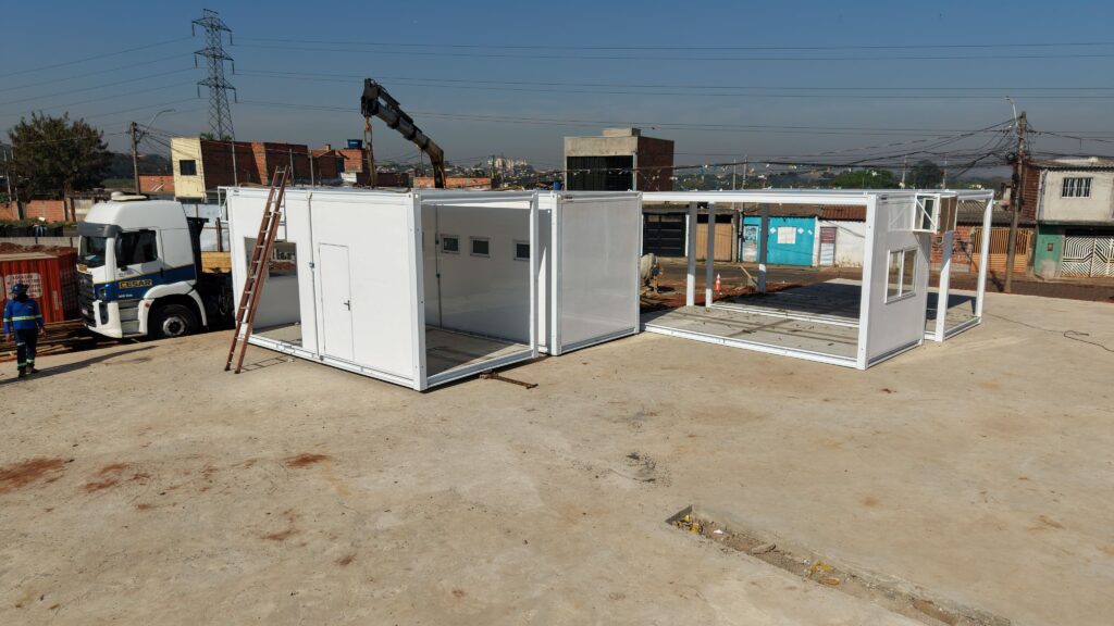 Esta imagem apresenta a montagem de estruturas modulares pré-fabricadas em um terreno aberto, com um caminhão equipado com guindaste realizando o posicionamento dos módulos brancos sob um céu limpo. A composição da cena é organizada de forma funcional e industrial, centrando-se no alinhamento de vários módulos habitacionais ou de escritório de cor branca. À esquerda, um caminhão branco está estacionado com um guindaste em operação, conectado à estrutura, enquanto uma escada de madeira está encostada em um dos blocos. A disposição dos módulos sugere uma fase ativa de construção ou montagem, com espaços abertos e estruturas metálicas expostas, ocupando a maior parte do plano médio da imagem em um cenário de terreno plano e batido. Os elementos principais incluem os módulos pré-fabricados, que são estruturas metálicas brancas de formato retangular, com design minimalista, janelas pequenas e portas de acesso. O caminhão, um veículo de carga pesado, apresenta cabine branca e detalhes em azul, servindo como suporte logístico para o içamento das peças. Um operário, visível no canto inferior esquerdo, veste uniforme azul de segurança, conferindo escala humana à construção. O guindaste, braço mecânico articulado de cor preta, é o ponto de conexão visual entre o caminhão e o módulo que está sendo posicionado. Esta é uma fotografia realista, capturada sob luz natural intensa, possivelmente durante a manhã ou início da tarde, o que minimiza sombras profundas e destaca a cor branca das estruturas contra o solo terroso. A execução técnica é clara, com um foco nítido em toda a cena, típica de registros de documentação de obra ou engenharia. O estilo é puramente documental e funcional, priorizando a clareza dos detalhes sobre a estética artística, utilizando uma perspectiva angular que permite visualizar a extensão total da montagem. O cenário é um pátio de construção em uma área semi-urbana, rodeado por edificações mais antigas e torres de transmissão de energia ao fundo. O tempo está ensolarado com um céu azul sem nuvens, criando uma iluminação ambiente brilhante e uniforme que preenche o espaço. O solo é composto de terra batida e cimento, reforçando a natureza industrial e pragmática do ambiente. A atmosfera é de um canteiro de obras em atividade, transmitindo uma sensação de progresso, organização logística e trabalho técnico eficiente sob condições climáticas ideais.