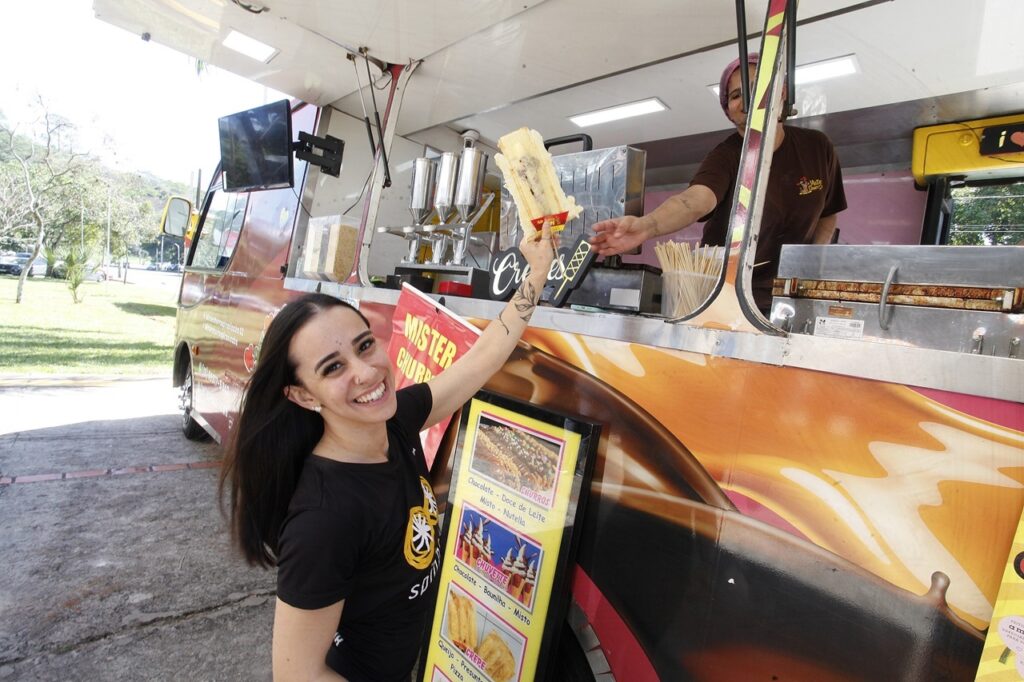 A imagem mostra uma cena alegre em um food truck especializado em churros e crepes. Em primeiro plano: Uma jovem sorridente está em pé, de frente para a câmera. Ela veste uma camiseta preta e tem uma tatuagem no braço levantado, com o qual ela segura um crepe preparado, entregando-o ou oferecendo-o para quem está dentro do trailer. No food truck: Há uma pessoa trabalhando dentro do trailer, vestindo uma camiseta marrom e touca, que estende a mão para receber ou completar a entrega do lanche. O trailer é adesivado com tons de marrom e laranja, simulando calda de chocolate ou doce de leite escorrendo. Menu: À frente, há uma placa com fotos e nomes dos produtos oferecidos, como "Churros" (sabores chocolate, doce de leite, misto, Nutella), "Chuvette" (chocolate, baunilha, misto) e "Crepe" (queijo, presunto, pizza). Ambiente: O cenário parece ser uma área externa, possivelmente um estacionamento ou parque, com gramado e árvores ao fundo sob a luz do dia. O ambiente transmite descontração e um atendimento simpático.