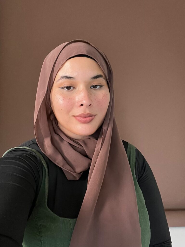 Esta imagem é um retrato de uma jovem mulher usando um hijab. Aqui estão os detalhes: A Pessoa: Ela tem uma expressão serena e um olhar direto para a câmera. Sua pele tem sardas naturais visíveis, e ela está usando uma maquiagem leve, destacando um delineado fino nos olhos e um tom natural nos lábios. O Hijab: Ela veste um hijab em um tom de marrom quente que emoldura seu rosto de forma elegante. Vestimenta: Por baixo do hijab, ela usa uma blusa preta de mangas compridas e um vestido ou avental em tom de verde musgo (provavelmente um tecido como veludo cotelê ou algodão grosso). O Ambiente: O fundo é um tom sólido de marrom, combinando harmoniosamente com as cores do hijab, criando uma composição limpa e focada nela. É uma foto muito bonita, com uma iluminação suave que valoriza bastante as feições dela!