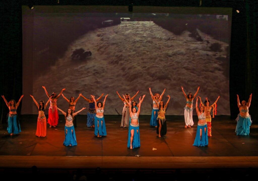 A imagem mostra um grupo de dançarinas apresentando uma coreografia em um palco. Elas usam figurinos coloridos, com saias longas, franjas, brilhos e acessórios típicos da dança oriental, principalmente em tons de azul, vermelho, dourado e prata. As bailarinas estão distribuídas pelo palco, com os braços levantados em diferentes poses. Ao fundo, um grande telão exibe uma imagem escura de água corrente ou de uma paisagem vista de cima. A iluminação alaranjada destaca o palco, enquanto a plateia aparece apenas como silhuetas na parte inferior.