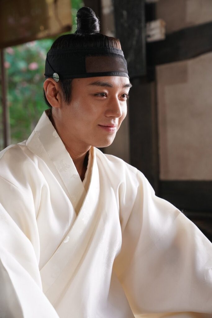 Esta imagem retrata um homem jovem de traços asiáticos vestindo um hanbok (traje tradicional coreano) branco, com um tecido que apresenta uma textura ou padrão sutil. Aqui estão os detalhes observados: O Traje: Ele veste a parte superior do traje em um tom de branco creme, com gola transpassada e um estilo clássico e elegante. O Penteado: O cabelo está preso no topo da cabeça em um coque, coberto por um manggeon (uma faixa de cabelo tradicional coreana feita de crina de cavalo, que era usada para prender os fios no lugar). A Expressão: Ele possui uma expressão serena e um leve sorriso, olhando para o lado, como se estivesse ouvindo ou conversando com alguém fora do enquadramento. Estilo: A iluminação é suave e focada, destacando o rosto e a textura da roupa, o que confere à imagem uma qualidade cinematográfica, típica de produções de época coreanas (os chamados sageuk).