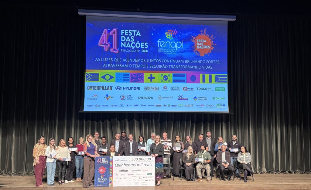 A imagem mostra um grande grupo de pessoas reunido no palco de um teatro, diante de cortinas escuras. Algumas estão sentadas e outras em pé, segurando certificados ou cartões. No centro, o grupo exibe um cheque simbólico de R$ 500.000,00, com a frase “Quinhentos mil reais”. Ao fundo, uma tela azul anuncia a 41ª Festa das Nações de Piracicaba, com o tema “O Ano das Luzes”, além de vários logotipos de patrocinadores.
