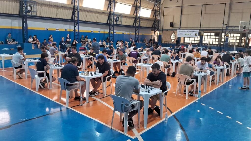 Uma competição de xadrez está acontecendo em um ginásio, com vários jogadores concentrados em suas partidas.
A imagem retrata um grande salão onde ocorre um torneio de xadrez, possivelmente dentro de um ginásio esportivo. O chão é dividido em áreas de laranja e azul, com linhas brancas demarcando o espaço. Dezenas de mesas brancas com conjuntos de xadrez estão dispostas em fileiras. Em cada mesa, indivíduos, em sua maioria jovens e adultos, estão sentados, concentrados em jogos de xadrez. Alguns jogadores estão inclinados sobre os tabuleiros, analisando os movimentos, enquanto outros parecem estar em contemplação. Há também espectadores sentados em arquibancadas improvisadas ou em cadeiras nas laterais, observando as partidas. A iluminação parece ser artificial, vinda de luminárias no teto, o que cria um ambiente claro e funcional. As paredes do ginásio são azuis e marrons, com algumas bandeiras e sinalizações penduradas. A atmosfera geral é de concentração e silêncio, com um tom de seriedade e competitividade sutil, típico de eventos de xadrez. As cores predominantes são o laranja e o azul do chão, o branco das mesas e os tons neutros das roupas das pessoas e da estrutura do ginásio.