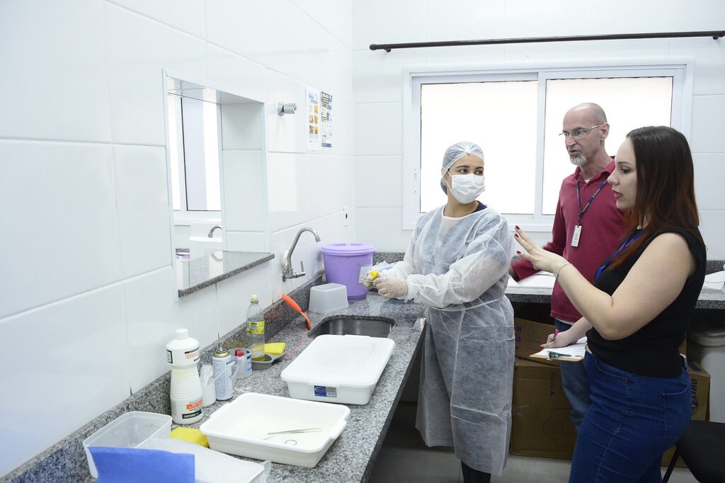 três pessoas estão em um ambiente clínico ou laboratorial. Uma delas veste equipamentos de proteção — touca, máscara e avental descartável — e se posiciona próxima a uma pia com bandejas, frascos e materiais de limpeza. As outras duas, uma com camiseta vermelha e outra de preto, parecem orientar ou discutir procedimentos com quem está paramentado.