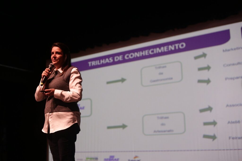 Na imagem há uma mulher em pé num palco falando ao público: Ela segura um microfone na mão direita e um controle/laser na esquerda. Veste camisa branca com suéter cinza por cima e calça escura. Ao fundo, projetada na tela, aparece um slide grande com o título em roxo “TRILHAS DE CONHECIMENTO” e vários blocos e setas verdes (fluxograma/organograma). Iluminação de palco foca a palestrante; o restante do palco e o teto estão escuros. A composição é em grande plano médio à esquerda, com a tela ocupando a direita da foto, dando ênfase tanto na pessoa quanto no conteúdo apresentado.