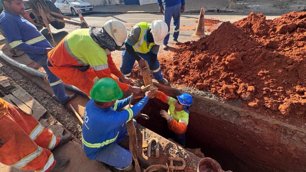 A imagem mostra uma cena de construção, possivelmente uma obra de saneamento ou tubulação, com vários trabalhadores interagindo em uma vala. No centro da imagem, há uma vala com paredes de terra avermelhada e compactada. Vários trabalhadores, todos usando equipamentos de segurança como capacetes e coletes de alta visibilidade, estão engajados em alguma tarefa dentro ou ao redor da vala. Os trabalhadores vestem roupas de trabalho coloridas, predominantemente em tons de laranja, azul e verde fluorescente. Alguns estão ajoelhados ou agachados, enquanto outros estão em pé. Há uma montanha de terra avermelhada à direita da vala, indicando que a escavação está em andamento. No fundo, ao longe, pode-se ver uma estrada asfaltada com um carro branco e cones de trânsito laranja, sugerindo que a obra está ocorrendo em uma área urbana ou em uma via pública. Um tubo metálico ou de plástico cinza claro está visível ao longo da parte superior da imagem, posicionado próximo à borda da vala. Há também alguns pedaços de metal enferrujado, possivelmente peças antigas de tubulação ou ferramentas, visíveis dentro da vala, em contato com a terra. A composição da imagem é dinâmica, com as figuras dos trabalhadores e os elementos da obra preenchendo a maior parte do quadro. A luz parece ser natural, possivelmente luz do dia, criando sombras e realçando as texturas da terra e das roupas. As cores predominantes são os tons terrosos da terra avermelhada, contrastando com as cores vibrantes dos uniformes de trabalho e os cinzas dos materiais. A perspectiva da câmera está ligeiramente inclinada, dando uma sensação de profundidade e imersão na cena de trabalho.