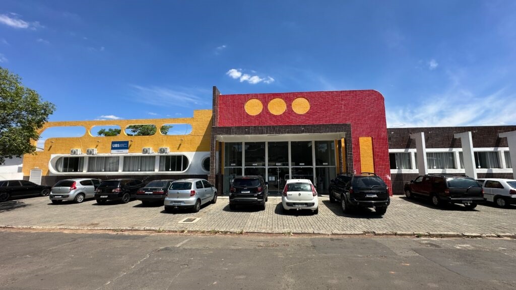 A imagem mostra a fachada de uma unidade de saúde identificada pela placa “UBS”. O prédio tem um design colorido, com revestimentos amarelo, vermelho e preto, além de três círculos amarelos na parte superior da entrada. Na frente, há uma área de estacionamento com vários carros. O céu está azul, com poucas nuvens, e uma árvore aparece no lado esquerdo da imagem.