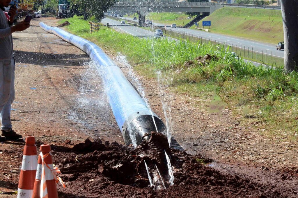A imagem mostra um grande tubo azul rompido em uma área de obras às margens de uma rodovia. A água jorra com força pela extremidade aberta, espalhando-se pelo solo de terra e formando poças. No primeiro plano, há cones de sinalização e montes de terra. Ao fundo, veículos trafegam pela rodovia, que passa sob um viaduto. Uma placa azul indica o “Viaduto Dr. João Rossi Salomão”. Na lateral esquerda, algumas pessoas observam ou registram o vazamento com celulares.