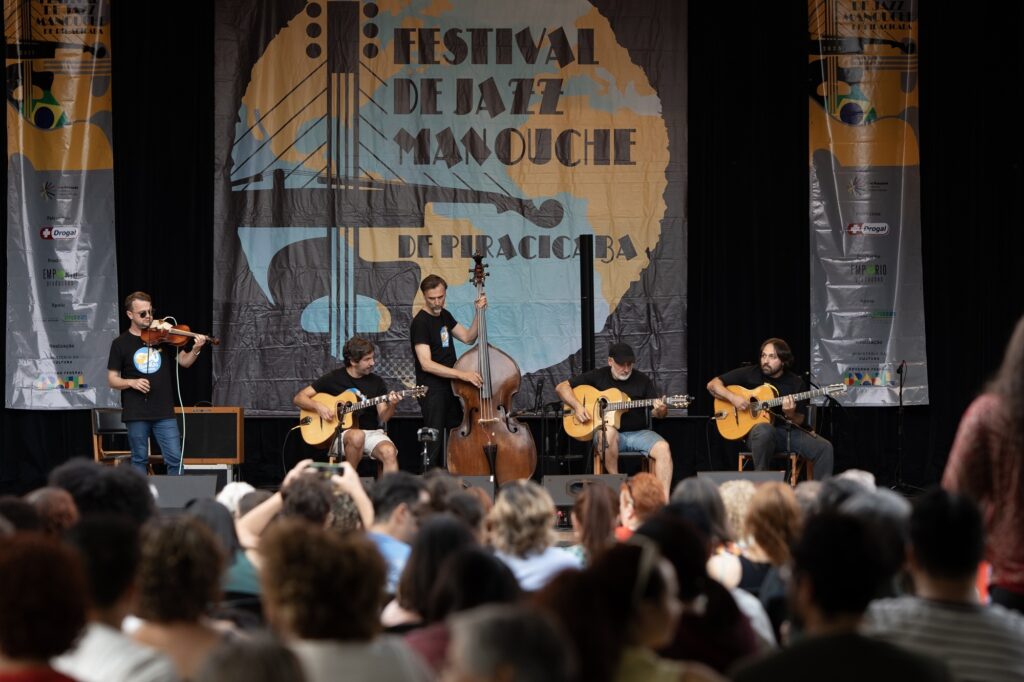 Esta imagem mostra um grupo de músicos no palco durante uma apresentação ao vivo no "Festival de Jazz Manouche de Piracicaba". O Grupo: Há cinco músicos no palco, todos vestidos de forma casual (camisetas pretas). À esquerda, um homem toca violino. Ao lado, um músico toca violão selmer-maccaferri (típico do Jazz Manouche). No centro, um contrabaixista toca um contrabaixo acústico grande. À direita, outros dois músicos tocam violões selmer-maccaferri. O Palco: Ao fundo, há um painel grande com o título do festival, ilustrado com a silhueta de um violão. Nas laterais do palco, há banners verticais com as marcas dos patrocinadores e apoiadores, incluindo logos da Lei Rouanet, Drogal, Empório Produções e Prefeitura de Piracicaba. A Plateia: Em primeiro plano, vemos a parte de trás da cabeça e os ombros das pessoas que compõem o público. Algumas pessoas na plateia estão segurando celulares, provavelmente gravando ou tirando fotos da apresentação. Atmosfera: O clima é de um evento cultural organizado e focado, com a plateia atenta ao concerto.