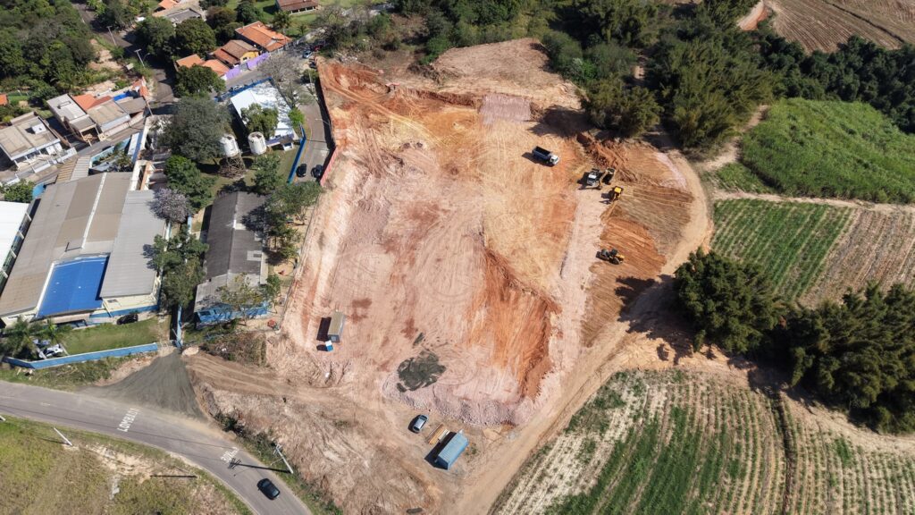 Essa imagem aérea mostra um terreno em obras, já preparado para receber construção, com solo avermelhado sendo movimentado por escavadeiras, tratores e caminhões. O espaço contrasta com o entorno: campos verdes, árvores e casas de telhados claros, uma delas com piscina azul visível. Na parte inferior, uma estrada pavimentada conecta o canteiro à vizinhança próxima. A