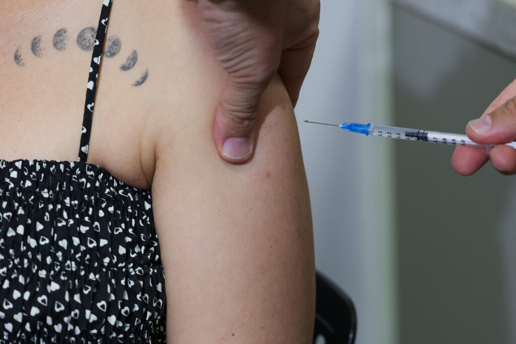 Esta imagem mostra um procedimento de vacinação ou aplicação de injeção. Aqui estão os detalhes: A Ação: Uma pessoa (profissional de saúde) está preparando ou aplicando uma injeção no braço de outra pessoa. A mão do profissional segura firmemente a pele do ombro/braço, enquanto a outra mão segura a seringa com a agulha posicionada próxima ao local da aplicação. Detalhes: A seringa é fina, com marcações de graduação visíveis no corpo plástico. O Contexto: A pessoa que recebe a aplicação veste uma blusa preta com estampas brancas de corações. É interessante notar que ela possui uma tatuagem nas costas, na altura do ombro, que retrata as fases da lua.