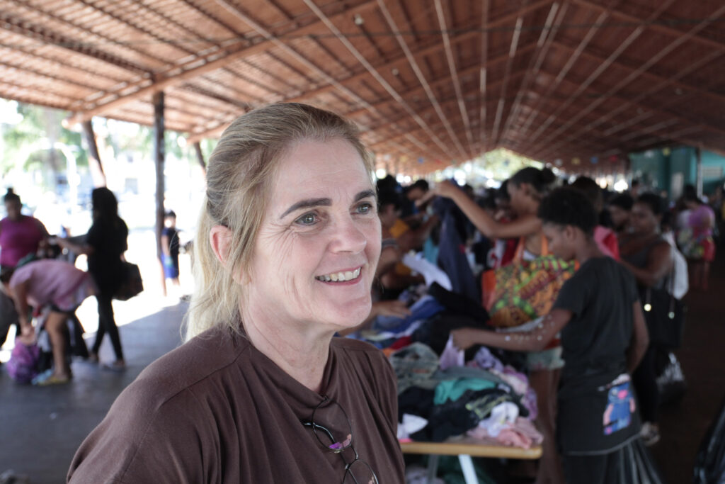 A imagem mostra uma mulher sorridente em primeiro plano, sob uma grande cobertura de madeira, enquanto várias pessoas ao fundo participam de uma ação comunitária de doação de roupas. As mesas estão cheias de peças de vestuário, e os participantes parecem escolher e organizar os itens. O ambiente é movimentado e iluminado pela luz natural, transmitindo um clima de solidariedade e engajamento social.