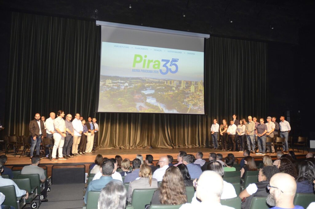 Uma fotografia de evento captura um grupo de pessoas em um palco durante uma apresentação institucional, com uma grande tela exibindo a marca "Pira 35" perante uma plateia atenta. A composição é estruturada em torno de um palco elevado no centro de um auditório, onde os participantes estão organizados em duas linhas laterais. O enquadramento é amplo, permitindo visualizar tanto os palestrantes no palco quanto a audiência sentada em poltronas dispostas em fileiras no plano inferior. A disposição dos elementos cria um foco central na grande tela projetada, que serve como pano de fundo para as pessoas em pé, estabelecendo uma hierarquia visual clara entre o conteúdo apresentado e os indivíduos presentes. Os sujeitos em destaque são diversos homens e mulheres, vestindo trajes que variam do casual ao social, incluindo camisas de botões, calças de sarja, blazers e blusas formais. Alguns estão de pé no palco, possivelmente aguardando a vez de falar ou participando de uma cerimônia, mantendo posturas eretas e expressões profissionais. Na plateia, os indivíduos apresentam diferentes penteados, roupas variadas em tons sóbrios e posições corporais que sugerem atenção ao que é exposto na tela, como se estivessem em uma conferência ou evento corporativo. A imagem é um registro fotográfico documental, com estilo que prioriza a clareza da informação e o registro fiel do evento. A execução técnica foca em manter uma profundidade de campo que abrange tanto os participantes próximos à câmera quanto aqueles sobre o palco. A iluminação é controlada, vindo predominantemente da tela de projeção e das luzes do teto do auditório, o que cria um contraste entre o brilho da apresentação e a penumbra mais acentuada no ambiente da plateia, destacando os pontos de interesse na cena. O cenário é um auditório convencional, caracterizado por cortinas escuras ao fundo, poltronas estofadas e uma atmosfera de ambiente de trabalho ou conferência. A iluminação é artificial, com o brilho da tela projetada dominando o ambiente e banhando os rostos das pessoas em uma luz suave. O ambiente transmite seriedade e foco, sugerindo um momento de planejamento estratégico, onde a luz projetada pela tela não apenas ilumina o palco, mas também define o humor do encontro, concentrando a atenção coletiva nos objetivos institucionais representados pelo nome "Pira 35" e a imagem da cidade ao fundo.