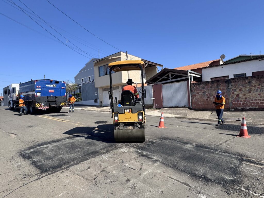 A imagem mostra uma equipe de trabalhadores fazendo manutenção no asfalto de uma rua residencial, sob um céu azul e ensolarado. No centro há um rolo compactador amarelo da Caterpillar, com um funcionário sentado na máquina. À esquerda, aparece um caminhão azul identificado como “UMCP 8”, enquanto outros trabalhadores usam uniformes laranja e equipamentos de segurança. Há cones de trânsito, áreas de asfalto recém-reparadas, casas ao redor e vários fios elétricos atravessando o céu.