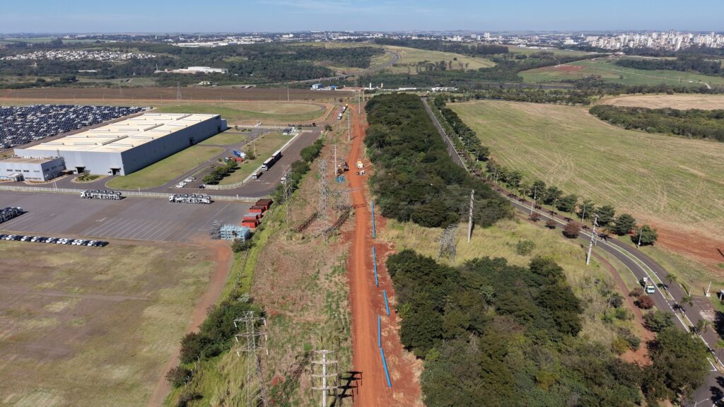 O imóvel é uma visão aérea de uma vasta área industrial com paisagens naturais e urbanas ao fundo. No primeiro plano, à esquerda, uma grande edificação industrial de cor cinza claro se destaca, com um telhado plano segmentado em branco. Ao lado da edificação, há um amplo estacionamento com diversas fileiras de carros e caminhões estacionados. Alguns caminhões transportadores de carros estão posicionados nas proximidades. Há também uma área de armazenamento com contentores vermelhos. O restante da área visível do primeiro plano é composto por campos com vegetação seca e um gramado baixo, intercalados com estruturas de alta tensão e seus cabos, característicos de uma rede elétrica. No centro da imagem, uma estrada de terra avermelhada se estende em direção ao fundo. Ao longo desta estrada, há trabalhos de construção em andamento, com máquinas de construção, pilhas de materiais e tubos azuis dispostos em intervalos regulares. A estrada está ladeada por uma área densa de vegetação de árvores verdes escuras e algumas áreas com grama seca. À direita da estrada de terra, estende-se um campo verde em formato de polígono, com sulcos visíveis indicando o plantio. Há uma estrada sinuosa com pavimento escuro que segue a topografia do terreno, margeada por árvores esparsas e postes de iluminação. Uma pequena estrada secundária com alguns postes de eletricidade também é visível. Alguns carros circulam nesta estrada. Ao fundo, a paisagem evolui para uma área mais densa de vegetação, seguida por uma área urbana com edifícios de diferentes tamanhos e cores, principalmente brancos e beges. A cidade se estende até o horizonte, sob um céu azul claro com poucas nuvens. A linha do horizonte é irregular devido à presença de montanhas e colinas suaves ao longe. As cores predominantes na imagem são os tons de verde da vegetação, o marrom da terra e dos campos secos, o cinza claro da edificação industrial, o branco dos carros e da cidade ao fundo, e o azul do céu. A composição da imagem é dividida diagonalmente, com a área industrial e de construção ocupando a maior parte do quadro, e a paisagem natural e urbana compondo o fundo. A luz solar é forte, indicando um dia claro e ensolarado, com sombras bem definidas, especialmente nas estruturas de alta tensão e na edificação. O ponto de vista é elevado, sugerindo uma captura feita por drone ou aeronave, proporcionando uma visão panorâmica detalhada da intersecção entre infraestrutura industrial, desenvolvimento urbano e paisagem natural.