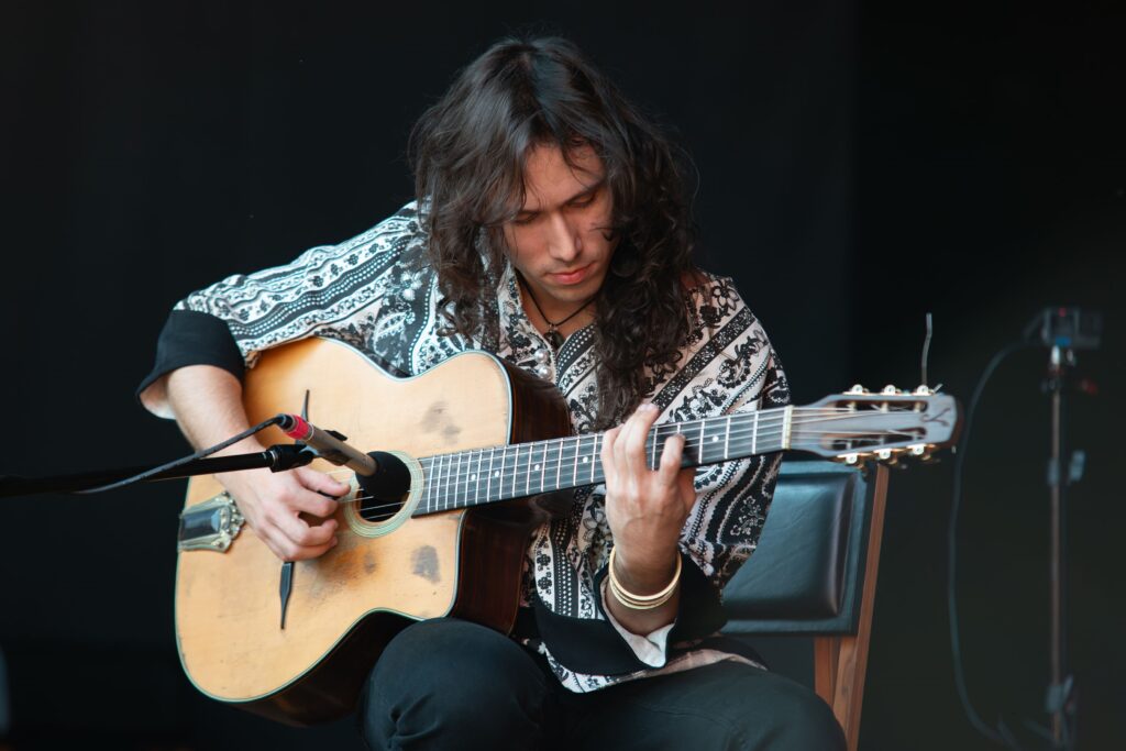 Esta imagem retrata um músico concentrado tocando um violão estilo gypsy jazz (também conhecido como violão cigano ou Manouche). Aqui estão os detalhes observados: O Músico: Ele tem cabelos longos e escuros, e veste uma camisa com uma estampa complexa em preto e branco (estilo paisley ou padrão étnico). Ele parece estar focado intensamente em sua performance, olhando para o braço do instrumento. O Instrumento: O violão é o modelo clássico desse gênero, com uma boca em formato de "D" (ou "D-hole"), que é característica dos violões Selmer-Maccaferri. Há um microfone posicionado estrategicamente em frente à boca do instrumento para captar o som. O Ambiente: O fundo é um painel preto simples, o que coloca todo o foco no músico. Ele está sentado em uma cadeira de encosto escuro. Estilo: A iluminação é suave e uniforme, destacando os detalhes do violão, a textura da camisa e as mãos do músico, criando uma atmosfera artística e intimista, típica de uma apresentação ao vivo.