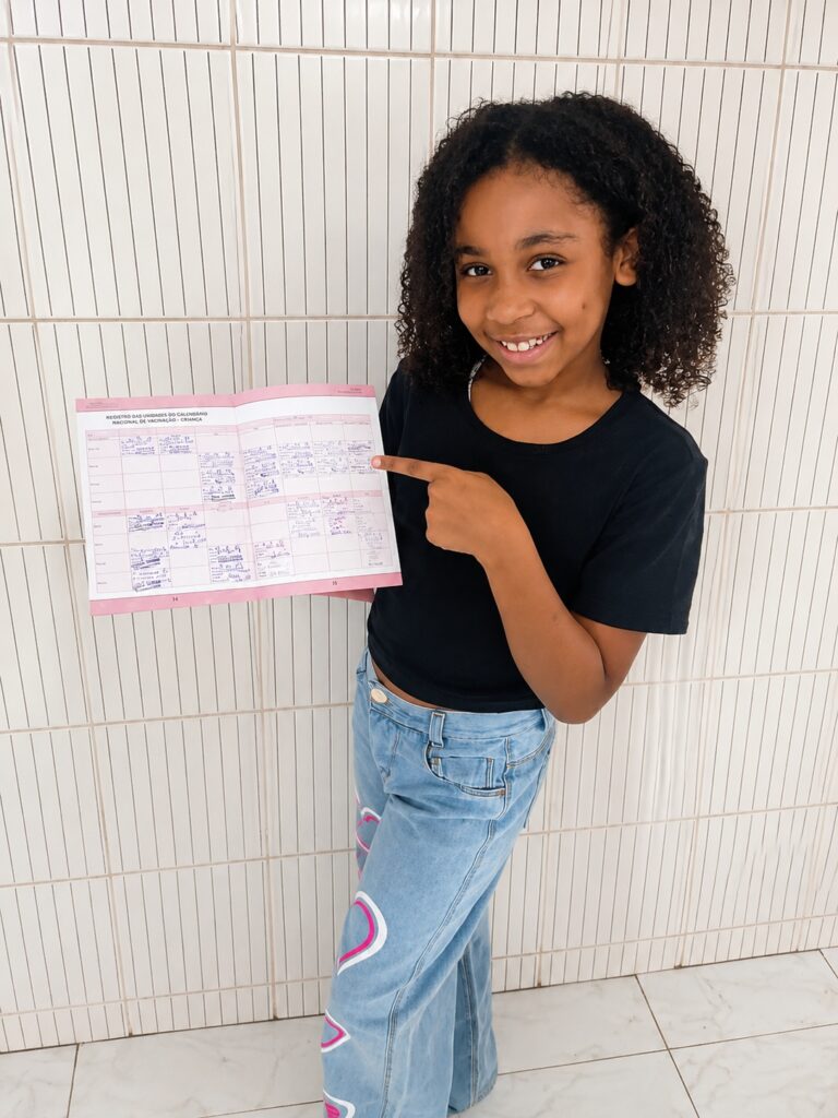 Uma pré-adolescente sorridente posa alegremente em frente a uma parede de azulejos claros, segurando e apontando para um livreto preenchido, transmitindo uma sensação de orgulho e realização por estar com a carteira de vacinação em dia. A composição é focada na menina, posicionada centralmente contra uma parede de azulejos verticais. Ela segura um livreto aberto com ambas as mãos, usando o dedo indicador da mão esquerda para apontar para o conteúdo, o que atrai o olhar do observador para a carteira de vacinação. A iluminação é uniforme e clara, destacando tanto a expressão feliz da jovem quanto os detalhes do livreto que ela apresenta. A perspectiva é frontal e ao nível dos olhos, criando uma conexão direta e amigável com quem vê a imagem. A menina tem cabelos escuros, cacheados e volumosos, com uma pele de tom quente e um sorriso aberto que revela alegria. Ela veste uma camiseta preta básica de mangas curtas e uma calça jeans clara, que apresenta detalhes decorativos em formato de coração nas laterais. Sua postura é relaxada e confiante, com o corpo levemente inclinado para o lado enquanto ela exibe o livreto. A imagem é uma fotografia digital clara e bem iluminada, capturada com foco nítido na pessoa e no objeto que ela sustenta. A qualidade técnica destaca texturas, como o padrão dos azulejos ao fundo e a trama do tecido das roupas. Não há manipulação artística pesada, mantendo o estilo de um registro fotográfico cotidiano que prioriza a autenticidade e a clareza do momento capturado. O uso de luz suave ajuda a evitar sombras duras, mantendo uma atmosfera limpa e agradável.