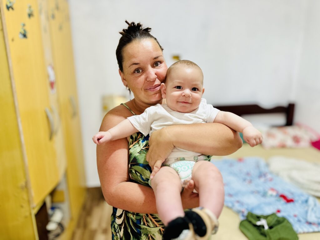 A imagem mostra uma mulher sorrindo enquanto segura um bebê no colo. O bebê, vestido de branco, olha diretamente para a câmera com uma expressão tranquila. Ao fundo, há um ambiente doméstico iluminado, com uma parede branca, um móvel ou armário amarelo e alguns objetos coloridos. A cena transmite carinho e proximidade entre mãe e filho.