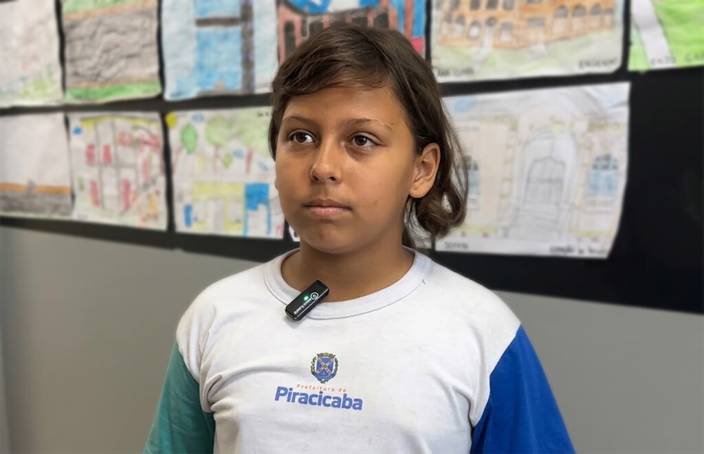 Nesta imagem, vemos um estudante jovem em ambiente escolar, vestindo uniforme branco e azul com o brasão e a inscrição “Prefeitura de Piracicaba”. Ao fundo, a parede está decorada com diversos desenhos coloridos, representando prédios, árvores e cenas urbanas, provavelmente produzidos por colegas, o que reforça o caráter criativo e coletivo da atividade.