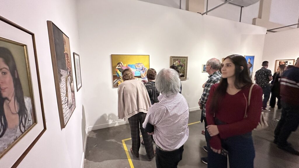 A imagem registra um momento de visitação em uma exposição de arte em galeria ou museu. O Público: Várias pessoas estão percorrendo o espaço, observando atentamente as obras expostas nas paredes brancas. Em destaque, à direita, uma jovem de blusa vinho observa as pinturas, enquanto outros visitantes, de costas ou de perfil, examinam as peças com interesse. As Obras: As paredes estão decoradas com quadros de diferentes estilos e tamanhos. Entre eles, percebe-se um quadro centralizado que retrata uma figura deitada sobre um móvel (sofá ou cama) em um ambiente amarelo, além de outras pinturas, incluindo retratos e naturezas-mortas. O Ambiente: O espaço é amplo, com teto alto, piso de cimento queimado demarcado com fitas amarelas de sinalização e iluminação focada nas obras, criando uma atmosfera cultural e organizada.