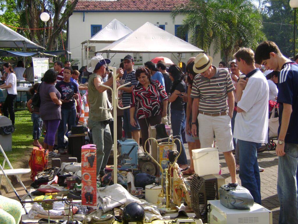 Na imagem, vemos um mercado ao ar livre, onde várias pessoas circulam entre mesas e bancas repletas de objetos de segunda mão, como eletrodomésticos, roupas, brinquedos e utensílios variados. O ambiente é descontraído e comunitário, com barracas montadas sob tendas e um prédio de telhado vermelho ao fundo, cercado por janelas azuis. A presença de palmeiras e vegetação reforça a sensação de clima quente e agradável. A cena transmite a ideia de um encontro popular voltado para compra, venda e troca de itens usados, destacando tanto a interação social quanto práticas de consumo mais sustentáveis.