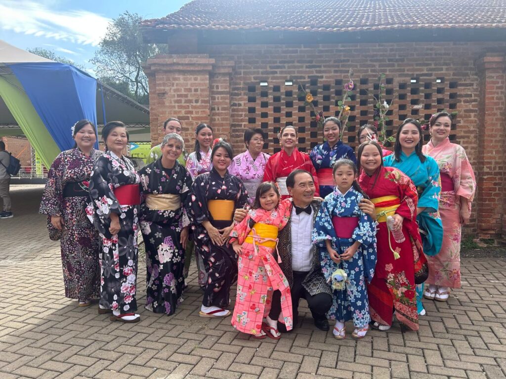 grupo maior, incluindo adultos e crianças, reunido para uma foto em área externa. Os participantes usam quimonos com estampas florais variadas, tendo ao fundo uma construção de tijolos e estruturas do evento.