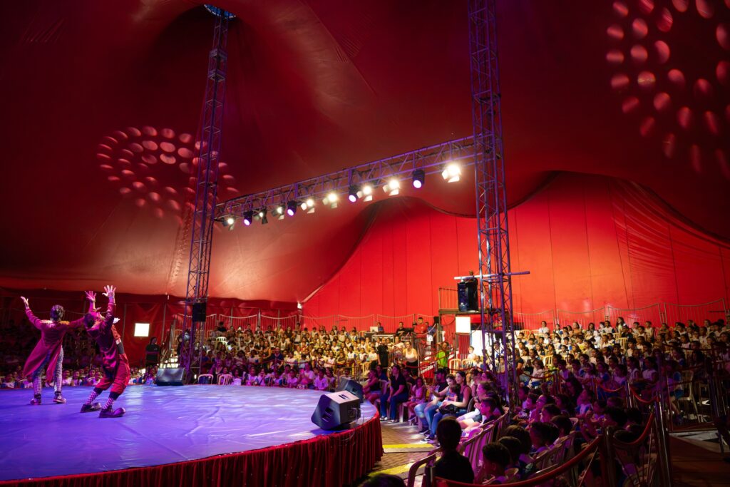 A imagem mostra um espetáculo circense dentro de uma grande tenda vermelha. No centro, há um palco circular iluminado por refletores coloridos, onde dois artistas — provavelmente palhaços — fazem uma apresentação animada. Em volta do palco, o público, composto por muitas crianças e adultos, assiste atentamente, criando uma atmosfera alegre e vibrante típica de um show de circo.