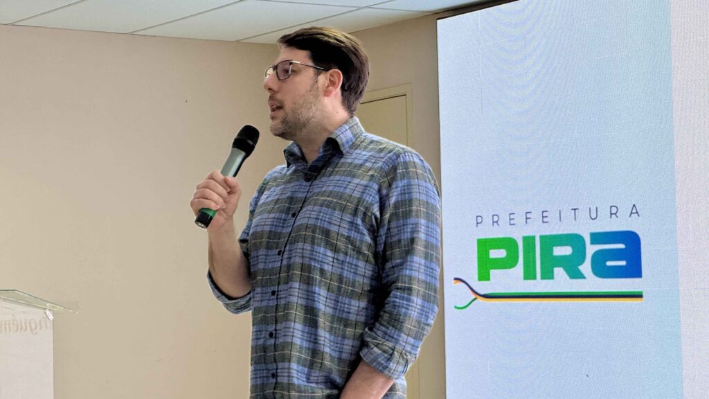 A imagem mostra um homem falando ao microfone durante uma apresentação em ambiente interno. Ele veste uma camisa xadrez azul e cinza e usa óculos.