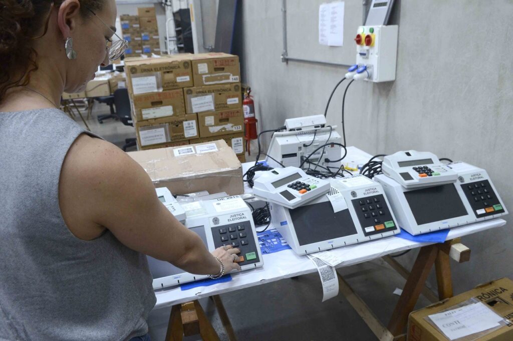 Uma funcionária de perfil manuseia e testa urnas eletrónicas dispostas sobre uma bancada no Cartório Eleitoral, com várias caixas de papelão empilhadas ao fundo.